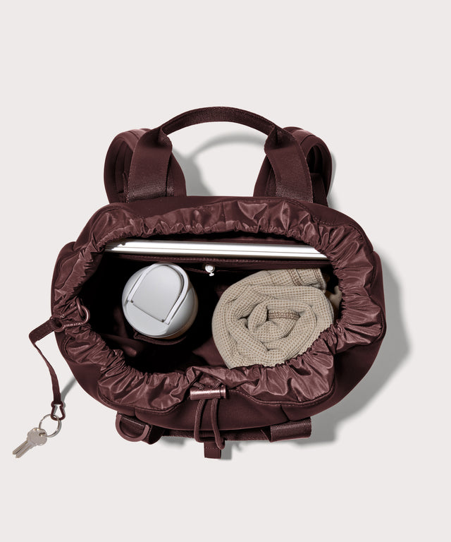Rhys Backpack in a deep brown with purple undertones unzipped, showing the interior pockets and drawstring closure.
